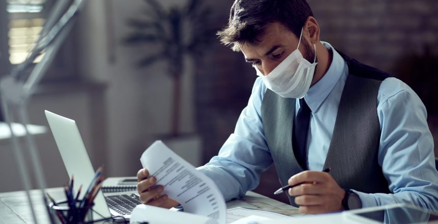 Male entrepreneur analyzing business reports while wearing face mask and working in the office during virus epidemic.