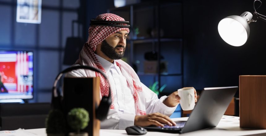 Side-view of a Muslim guy holding a cup of coffee and surfing the net on his digital laptop. Image showcasing a male Arab person engrossed in his personal computer while drinking tea.